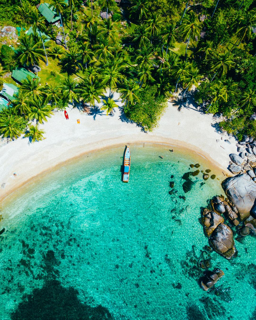 Aerial view of a quiet Koh Samui cove with turquoise water, white sand, a moored longtail and coconut palms