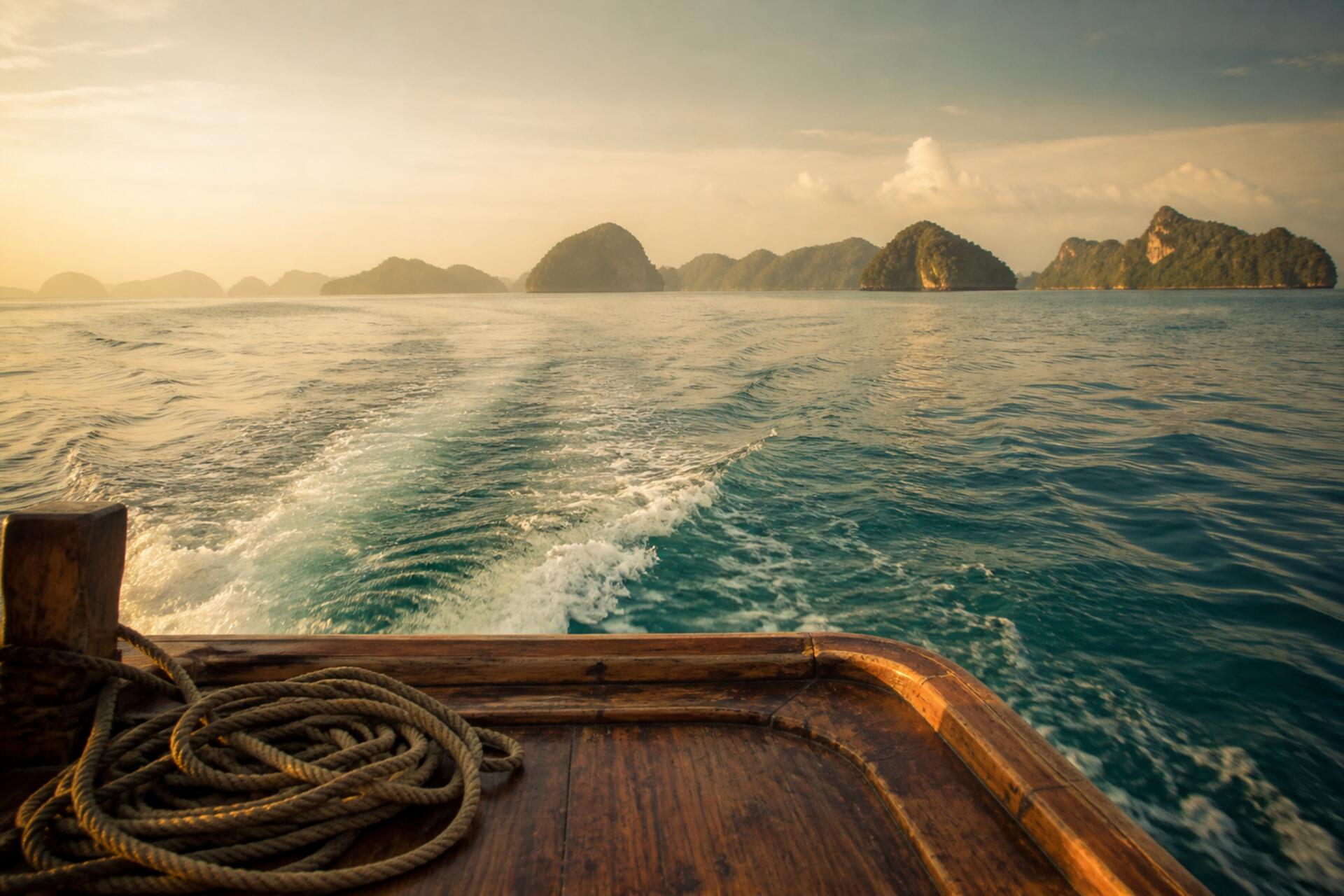 The view back from a wooden boat crossing calm water in Thailand, with a white wake behind and green islands in morning haze on the horizon